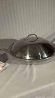 Photo of stainless steel wok with lid, showing profile with handle and lid handle, on white background.
