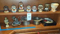 Shelf displaying nine assorted ceramic character ashtrays including faces with open mouths, a skull design, and one white marbled piece. Also includes two metal top hat-shaped ashtrays and a black ashtray holding gambling chips.