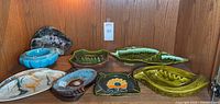 Full view of nine assorted ceramic ashtrays in various shapes and colors displayed on wood shelf.