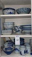 Full view of two shelves with blue and white patterned bowls and trays, small bowls and rectangular plates, teapot and sake cups on two shelves.