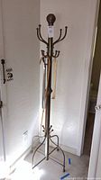 Full view of brass tone metal hat rack standing against wall, showing the height and base structure with caster wheels, and hanging items.