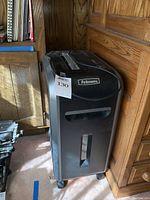 Front view of large black Fellowes paper shredder near wooden furniture, showing unit on caster wheels with a pull-out bin and small window.