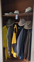 Closet view with yellow rain suit, multiple jackets, and several tan and beige hats hanging on top shelf and hooks