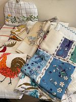 Overview of embroidered tea cozy alongside stack of printed tablecloths showing rooster motif and various floral prints