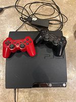 Black Sony Playstation 3 Slim console with two wireless controllers: one black, one red, resting on top, along with the power adapter cords.