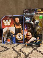 Photo showing both WWE action figures in sealed packaging on carpet: Braun Strowman on left, Zeus on right with accessories