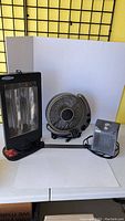 Three items side by side on table: one black rectangular heater, one circular black fan, and one small grey heater.