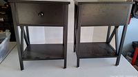 Front view of two black wooden end tables with one drawer, lower shelf, and x-shaped side supports.