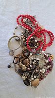 Heap of assorted used costume jewelry including red beaded necklaces, mixed metal chains, pendants, and bracelets on a light fabric background.