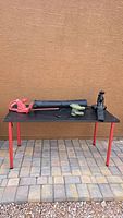 Wide shot of three corded electric yard tools: red Toro hedge trimmer, green Homesite trimmer, and black Homdox blower on black table outdoors.