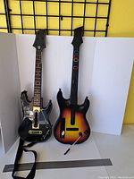 Two Guitar Hero style wireless guitar controllers including one black World Tour style and one sunburst color with fret buttons and whammy bar.