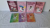 Seven children's paperback books on a white surface, including three 'Rainbow Magic' fairy books with colorful covers and four pink unicorn-themed books.