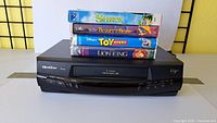 Front view of Quasar VHQ-940 VHS player with four VHS movies stacked on top: Shrek, Beauty and the Beast, Toy Story, and The Lion King.