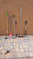 Six yard tools lined up against a wall on a tiled floor showing various hand tools and broom