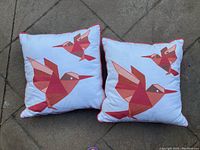 Photo showing two square decorative pillows with orange and white color tones and hummingbird designs placed on a concrete surface.