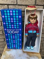 Full view of the American Girl doll in box with packaging visible, showing doll dressed in jacket, jeans, sneakers, and straw hat.