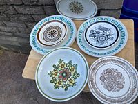 Shows all 12 dishes arranged on a small table outdoors against a stone wall. Includes varied styles and sizes, visible floral and geometric mid century patterns.