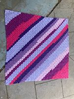 Full view of the crocheted lap blanket laid flat showing diagonal stripes in purple, pink, and lavender hues with textured crochet stitches.