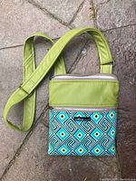 Crossbody purse with lime green adjustable strap and upper section, front zippered pocket with blue and green geometric pattern, placed on stone ground.