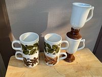 Front view of six stacked mugs, showing two white ceramic Bilton mugs with green and brown leaf patterns, and four other white mugs with brown bases.