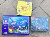 Lot showing three items: The Games for One Pack yellow box, Ocean View 1000pc puzzle with ocean life cover art, Women in Science 500pc puzzle.