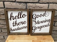 Photo of two wood signs side by side. Left sign says 'hello there handsome'; right sign says 'Good Morning Gorgeous.' Both have natural wood frames and white backgrounds with brown cursive text.