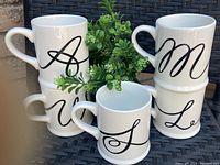 Set of six white ceramic mugs displayed on a dark woven surface with green foliage background, each mug featuring a large black scripted initial (A, M, W, L, S, U).