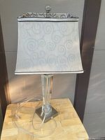 Full view of the table lamp showing the swirl patterned fabric lamp shade with silver trim and the clear and silver base.