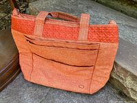 Photo showing the orange Lug tote purse from an angle, highlighting front zippered pockets and two shoulder straps.