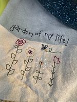 Close-up of beige kitchen tea towel with embroidery reading 'garden of my life' and colorful flowers