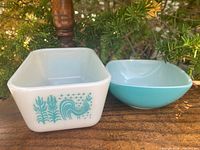 Rectangular white glass Butterprint fridgie with turquoise rooster and tulips alongside a solid turquoise square bowl on a wooden surface outdoors.