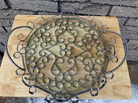 Front angle view of a round silver-tone metal serving tray with ornamental scrollwork, twisted handles, and removable clear glass insert on a wooden surface.