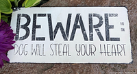 Photo of a rectangular tin sign with the text 'BEWARE: DOG WILL STEAL YOUR HEART' in black lettering on a white background, showing some distressed, rustic wear around the edges.