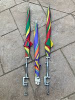 Three folded colorful patio umbrellas with metal clamp bases on a stone-paved outdoor surface.