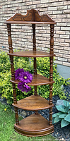 Full view of vintage wooden corner what not shelf with four triangular shelves and decorative top, set against an outdoor brick background with flowers.
