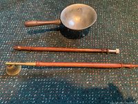 Full view of brass melting pot with wooden handle alongside a wooden-handled brass candle snuffer on fabric background.