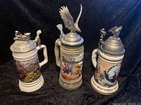 Three German beer steins arranged side by side, showing detailed artwork and pewter lids with finials.