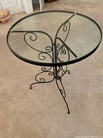 Full view of round glass top on black metal scrollwork base, placed on carpeted floor.