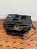 Full view of HP ENVY 7640 black all-in-one printer on wooden floor with power cord laid out.