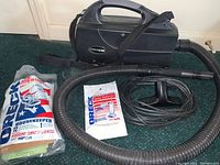 Photo of black Oreck Housekeeper canister vacuum with attached flexible hose, wide floor nozzle, power cord, and vacuum bags in original packaging on green carpet.