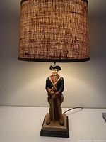 Lamp showing ceramic Revolutionary War soldier figurine as base with musket, detailed uniform, human face, and brown textured fabric lampshade casting warm light.
