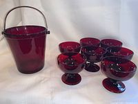 Ruby red glass ice bucket with metal handle next to seven parfait glasses arranged in an arc.