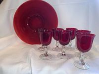 Ruby red starburst platter with seven matching stemmed glasses arranged in front, white fabric background.