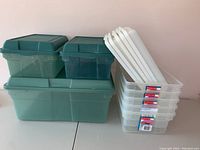 Group of stacked storage bins showing 2 medium green bins on top of 1 large green bin, next to 5 clear plastic bins stacked upright