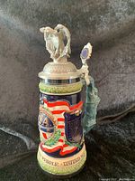 Full view of decorative beer stein with stars and stripes design and pewter eagle lid with figurine on top.