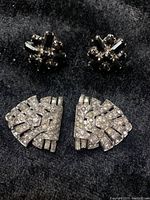 Pair of dark rhinestone Weiss screw back earrings next to two Art Deco style clear rhinestone scarf or fur clips on black velvet background.
