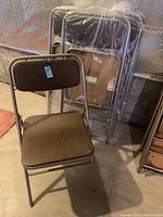 View of one vintage Samsonite folding chair unfolded, showing faux wood grain seat and back with bronze metal frame. Three other chairs are folded and wrapped in plastic behind.