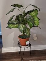 Full view of artificial plant in terracotta pot on black metal stand against wall.