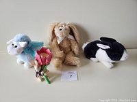Four Easter-themed decorations displayed together on a shelf: blue plush lamb, beige plush bunny, black and white plush rabbit, ceramic rabbit holding a red tulip