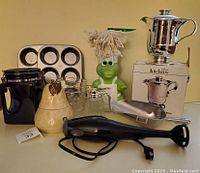 Photo showing the complete kitchen lot including hand blender, muffin tin, gravy boat, ceramic pear, black pitcher, glass shaker, metal pot, and alligator cleaning brush.